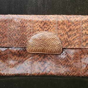 Peut inclure: Une pochette rectangulaire avec fermeture à rabat, en peau de serpent marron et beige. La pochette présente une surface texturée avec un motif d'écailles et un rabat arrondi sécurisé par un fermoir en peau de serpent assorti. Le design général est de tons naturels et terreux.