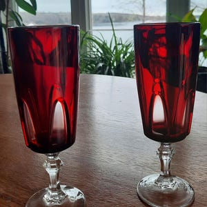 May include: Two red glass goblets with clear stems and bases. The goblets have a faceted design and are sitting on a wooden table.