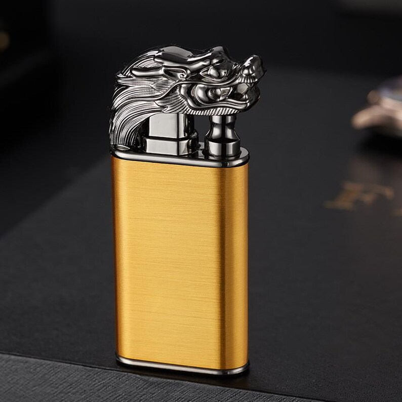 May include: A gold-colored lighter with a dragon head design. The lighter has a brushed metal finish and a black trim.