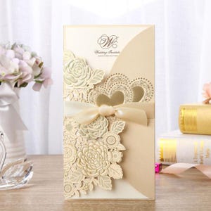 May include: A cream-colored wedding invitation card featuring an intricate laser-cut floral overlay on the left side, a smooth diagonal pocket, and a central champagne-colored satin ribbon bow. The text reads Wedding Invitation.