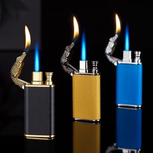 May include: Three butane lighters with alligator head designs. The lighters are black, gold, and blue. Each lighter has a double flame.