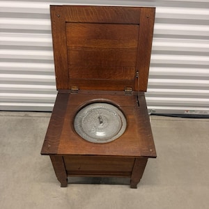 May include: A square wooden commode chair with a dark brown finish and a hinged lid that opens upward. The seat features a circular cutout containing a removable, circular metal basin with a central handle, set into the wooden frame.