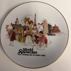 May include: A white ceramic decorative plate with a gold-toned rim featuring a colorful illustration of global landmarks and figures in traditional attire. The text reads World Showcase OPENING DAY OCTOBER 1, 1982 in black lettering at the bottom.
