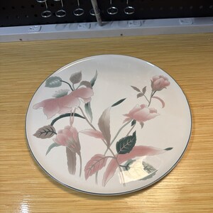 May include: A round ceramic dinner plate with a cream-colored base featuring a floral pattern of soft pink roses and muted green leaves. The plate has a thin, dark-colored rim around the edge, providing a clean and simple border to the design.