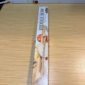 May include: A rectangular cardboard product box for The Original Fiddle Bow Bread Knife. The packaging features a graphic illustration of a wooden bread knife slicing through a loaf of bread and a tomato, with the product name printed in black text.