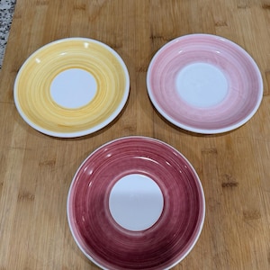 May include: Three ceramic saucers in yellow, pink, and burgundy, each with a white center and a painted spiral design. The saucers are arranged on a wooden surface, showcasing their colorful, decorative patterns.