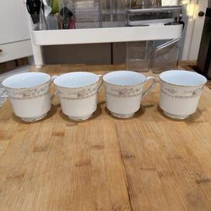 May include: Four white teacups with gold rims and floral patterns. The cups have handles and are arranged on a wooden surface. The floral design includes pink and gold accents, adding a touch of elegance.