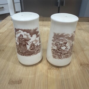 May include: A pair of white ceramic salt and pepper shakers with brown transferware designs. One shaker depicts a couple, the other a person holding a flag that reads "DON'T GIVE UP THE SHIP".