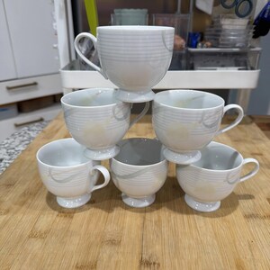 May include: Six white teacups stacked on a wooden surface. Each cup has a pedestal base, a curved handle, and a decorative gray swirl and line pattern. The cups are arranged in a pyramid shape.