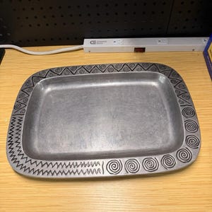 May include: A rectangular silver-toned metal tray featuring a wide, flat border decorated with engraved black patterns. The border design includes a repeating sequence of spirals, triangles, and zigzag lines etched into the metallic surface.