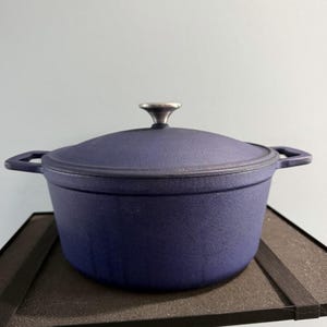 May include: A navy blue cast iron Dutch oven with a lid and side handles. The lid has a silver-colored knob. The pot is sitting on a black surface.