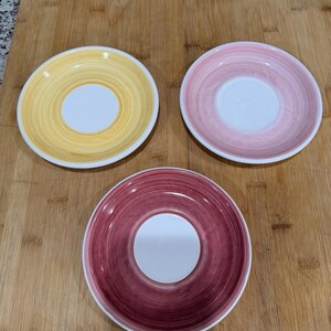 May include: Three round ceramic saucers with a white center and a wide, hand-painted rim featuring a swirling brushstroke pattern. One saucer has a yellow rim, one has a light pink rim, and the third has a deep magenta rim.