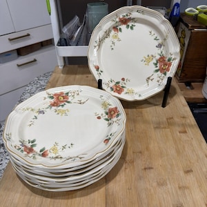 May include: A stack of off-white ceramic plates with a scalloped edge and floral design. The plates feature red, yellow, and green flowers with a gold trim. One plate is displayed on a black stand. The plates are on a wooden surface.