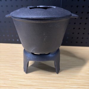 May include: A black cast iron fondue pot with a matching lid and a sturdy three-legged base. The pot features a wide, tapered bowl shape with small side handles, designed to sit securely on the integrated stand for heating or serving purposes.