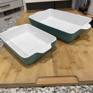 May include: Two rectangular baking dishes. The dishes have a white interior and a teal exterior. The dishes are sitting on a wooden cutting board. The cutting board is on a granite countertop.