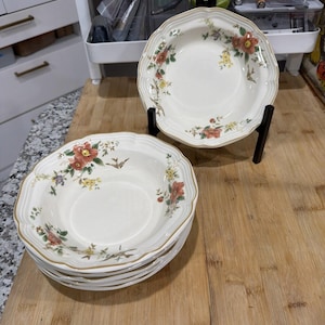 May include: A set of white ceramic bowls with a floral design featuring red, yellow, and green flowers. The bowls have a scalloped edge and a gold trim. One bowl is displayed on a black stand, while the others are stacked on a wooden surface.