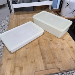 May include: Two rectangular, off-white plastic food storage containers with lids are on a light brown wooden cutting board. The containers are empty and appear clean. The cutting board has a handle on one side.