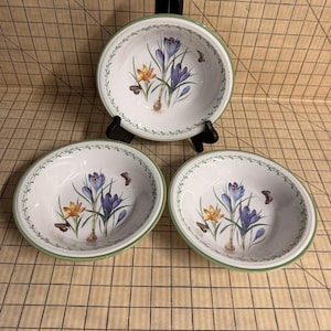 May include: Three white ceramic bowls featuring a botanical illustration of purple and yellow crocus flowers with small brown butterflies. Each bowl has a green rim and a delicate green vine border pattern around the interior edge of the basin.