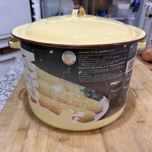 May include: A yellow enamel corn pot with a lid and side handles. The pot has a brown rim and a label with text and an image of corn on the cob. The pot is designed for boiling corn and stews.