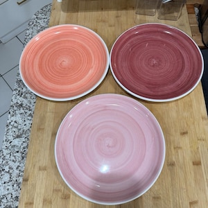 May include: Three ceramic plates with a spiral design. The plates are in shades of coral, burgundy, and pink, each with a white rim. They are arranged on a wooden cutting board, showcasing a simple, elegant aesthetic.