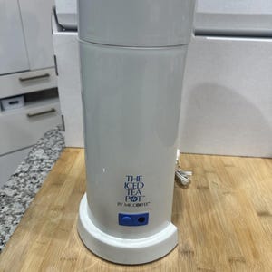 May include: White electric iced tea maker with a cylindrical design. The text "THE ICED TEA POT" is printed in blue. It has a blue button and a black button. The tea maker is on a wooden surface.