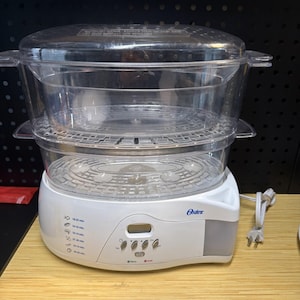 May include: A white Oster electric food steamer with a clear plastic two-tier steaming basket and lid. The base features a digital display, control buttons, and printed cooking time icons for various food items, along with a white power cord.