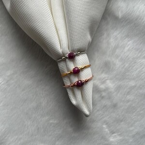 May include: Three wire-wrapped rings with oval, deep red gemstones. The rings are silver, gold, and rose gold in colour. The rings are displayed on a white, textured fabric, with a soft, white background.