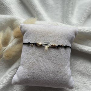 Pode incluir: Uma pulseira delicada com uma pedra clara e arredondada no centro, acentuada com contas douradas e pequenos detalhes. A pulseira é presa em um cordão preto, apresentada em uma almofada cinza sobre um fundo branco macio.