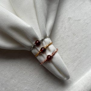 May include: Three wire-wrapped rings, each with a small, round, red gemstone. The rings are silver, gold, and copper-coloured, and are displayed on a white, textured fabric.