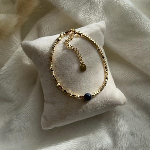 Può includere: Un braccialetto dorato con piccoli collegamenti rettangolari e una singola perla blu scuro. Il braccialetto è appoggiato su un cuscino bianco.