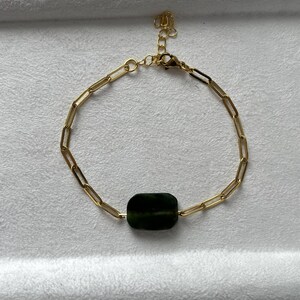 Könnte beinhalten: Goldfarbenes Kettenarmband mit einem dunkelgrünen, rechteckigen Edelstein. Das Armband hat ein filigranes Büroklammer-Kettendesign und einen Karabinerverschluss. Der Edelstein ist der Blickfang und verleiht dem minimalistischen Design einen Farbtupfer.