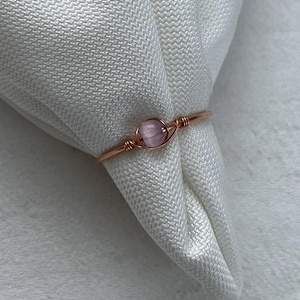 May include: A delicate ring featuring a small, round, pink gemstone set in a copper-colored wire. The ring is displayed on a white, textured fabric, highlighting the contrast between the metal and the stone. A simple, elegant piece of jewelry.