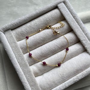 May include: A delicate gold bracelet featuring small, round, deep red gemstones. The bracelet has a fine chain and a simple clasp. The gemstones are evenly spaced along the chain. The bracelet is displayed on a white, textured surface.