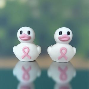 May include: Two white rubber duck figurines with pink beaks and a pink ribbon on their chests. The ducks have black eyes and are reflected in the surface below. The background is a blurred green.