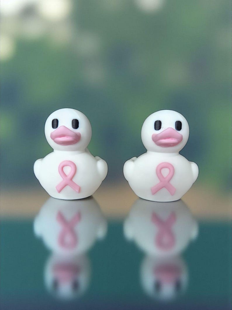 May include: Two white rubber duck figurines with pink beaks and a pink ribbon on their chests. The ducks have black eyes and are reflected in the surface below. The background is a blurred green.
