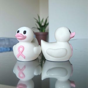 May include: Two white rubber duck toys with pink beaks and eyes. One duck has a pink ribbon on its chest. The ducks are on a reflective surface, with a plant in the background. The ducks are a symbol of breast cancer awareness.