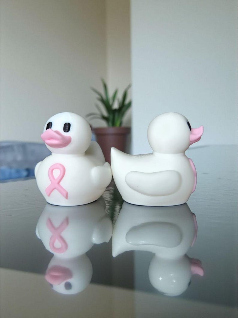 May include: Two white rubber duck toys with pink beaks and eyes. One duck has a pink ribbon on its chest. The ducks are on a reflective surface, with a plant in the background. The ducks are a symbol of breast cancer awareness.