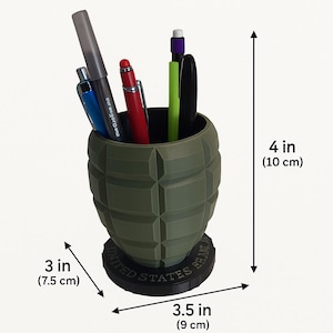 May include: A green desk organizer shaped like a grenade, 4 inches (10 cm) tall and 3.5 inches (9 cm) wide, holding pens and a marker. The base reads "UNITED STATES BRANC". The organizer contains a blue pen, a red pen, a green pen, a black pen and a gray marker.
