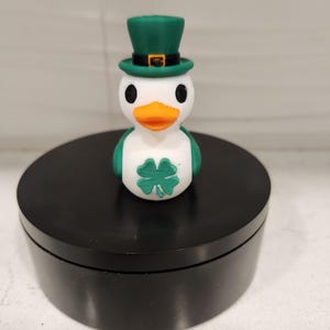 May include: A white and green duck figurine wearing a green top hat with a gold buckle. The duck has an orange beak, black eyes, and a green shamrock on its chest. The figurine is on a black circular stand.