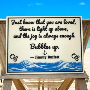 May include: A white sign with a blue wave design at the bottom. The sign has a quote by Jimmy Buffett that reads "Just know that you are loved, there is light up above, and the joy is always enough. Bubbles up."