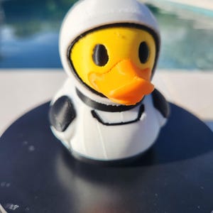 May include: A yellow rubber duck wearing a white astronaut helmet and spacesuit with black accents. The duck has an orange beak and black eyes. The duck is sitting on a black surface, with a pool in the background.