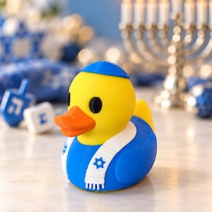 May include: A yellow rubber duck wearing a blue yarmulke and a blue-and-white scarf featuring a Star of David pattern. The duck has a bright orange beak and black eyes, with a blue body that contrasts against its yellow head and neck.
