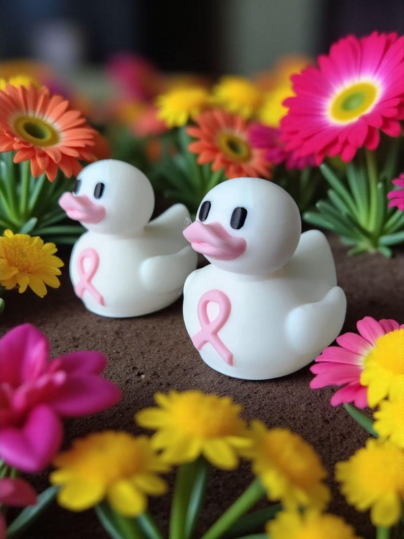May include: Two white duck figurines with pink beaks and a pink ribbon, surrounded by colorful flowers. The ducks have black eyes and are set against a blurred background of orange, yellow, and pink flowers.