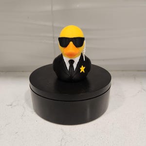 May include: A yellow rubber duck wearing black sunglasses, a black suit, and a tie. The duck has an orange beak and a gold star on its lapel. It is standing on a black, round platform.