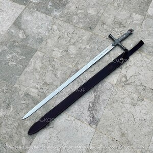 May include: A silver sword with an ornate handle and a long, straight blade, resting in a dark brown leather sheath. The sword's handle features intricate designs, and the blade has text etched along its length. The sheath is closed.