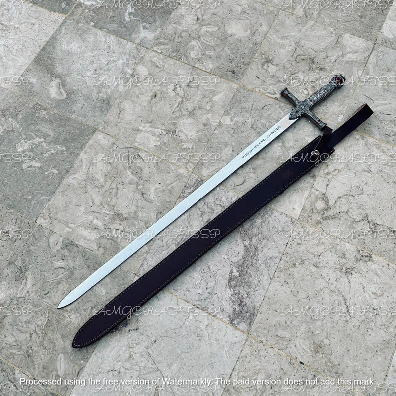 May include: A silver sword with an ornate handle and a long, straight blade, resting in a dark brown leather sheath. The sword's handle features intricate designs, and the blade has text etched along its length. The sheath is closed.