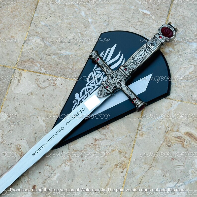 May include: A decorative sword with an ornate silver handle and a polished blade, resting on a black shield-shaped plaque. The handle features red accents and intricate carvings. The blade has the text "GODRIC GRYFFINDOR" engraved on it.