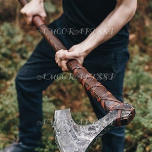 May include: A handcrafted axe with a wooden handle and a metal blade. The handle is wrapped with leather straps. The axe head has a textured, weathered appearance. The axe is held by a person wearing dark clothing.