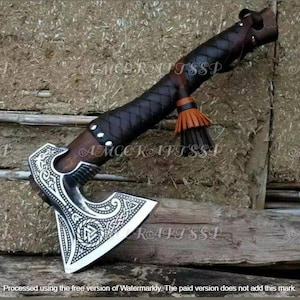 May include: A handcrafted axe with a silver blade featuring intricate designs and a wooden handle wrapped in black leather. The handle has a leather strap and a tassel. The axe is displayed on a wooden surface.