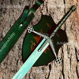 May include: A silver-toned sword with an ornate crossguard featuring four-circle ends, paired with a dark green leather scabbard and a dark green shield-shaped plaque displaying a partial orange dragon graphic and the text FIRE AND BLOOD.